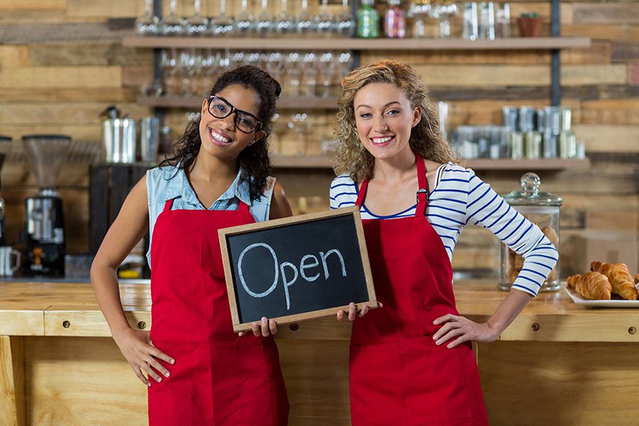 Business Insurance - Portrait of Two Smiling Young Female Business Owners Holding an Open Sign Together While Standing in Their Cafe